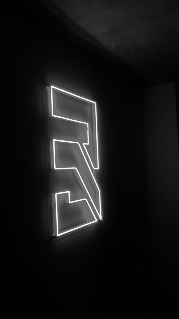 Het abstracte logo van Roliskos Barbershop (bestaande uit R en B samengevoegd) als neon lamp aan de muur